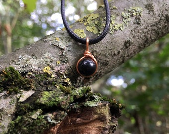 May include: A black gemstone pendant necklace with a copper wire wrap. The necklace is hanging from a tree branch.