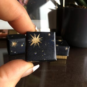 Può includere: Piccole scatole quadrate blu navy con un design celeste. Ogni scatola presenta un'esplosione di stelle dorate e minuscoli accenti scintillanti, che ricordano un cielo notturno. Le scatole sono tenute dalle dita di una persona.