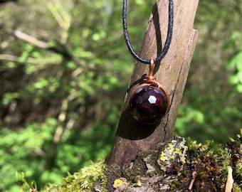 May include: A dark red gemstone pendant wrapped in copper wire, hanging from a black cord. The pendant is on a tree branch covered in moss.