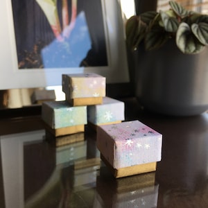 Peut inclure: Quatre petites boîtes carrées empilées les unes sur les autres. Les boîtes sont décorées d'un motif aquarelle rose pastel, bleu et violet avec des étoiles argentées scintillantes. Les boîtes ont une bordure dorée.