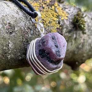May include: A raw, deep purple-red ruby crystal pendant wrapped in silver-toned wire. The stone features dark, textured inclusions and is suspended from a simple black cord necklace, creating a contrast between the metallic wire and the stone.