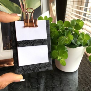 May include: A black faux leather notepad with a gold binder clip. The notepad has two white pages with a black line separating them. The notepad is sitting on a black surface.