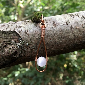 May include: A white gemstone pendant hangs from a copper wire wrapped loop. The pendant is suspended from a branch with a blurred green background.