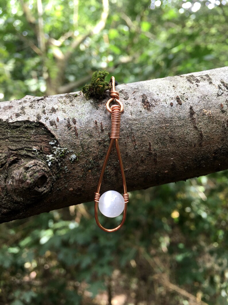 May include: A white gemstone pendant hangs from a copper wire wrapped loop. The pendant is suspended from a branch with a blurred green background.