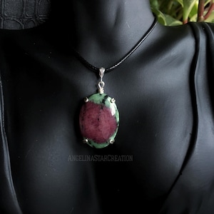 May include: A pendant necklace featuring a large, oval-shaped gemstone with a deep red center and green edges. The gemstone is suspended from a black cord with a silver-colored clasp. The necklace is displayed on a black mannequin.