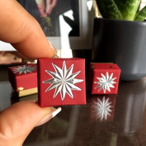 May include: A small, red gift box with a silver starburst design. The box is held between fingers, with two other boxes visible in the background. The text "ANGELINASTARCREA" is printed on the side of the box.