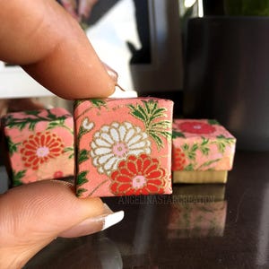 May include: Miniature square gift boxes with floral patterns in pink, white, red, and gold. The boxes are held by a person's fingers. The boxes have a decorative floral design. The boxes are small and appear to be handmade.