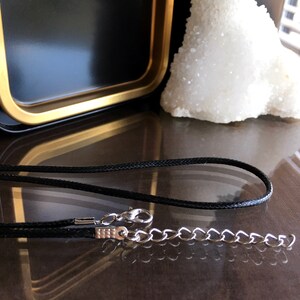 May include: A black cord necklace with a silver chain and clasp. The necklace is lying on a dark brown surface.