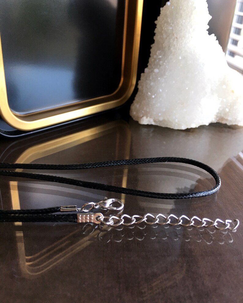 May include: A black cord necklace with a silver chain and clasp. The necklace is lying on a dark brown surface.