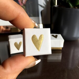 May include: A small, white square box with a gold heart design. The box is held between fingers, with two other similar boxes visible in the background. The boxes have a gold base and a gold heart on the front.