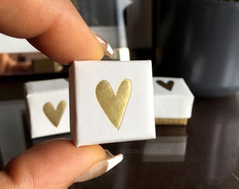 May include: A small, white square box with a gold heart design. The box is held between fingers, with two other similar boxes visible in the background. The boxes have a gold base and a gold heart on the front.