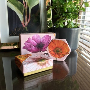 May include: Three small gift boxes with floral designs. The boxes are pink, orange, and white with floral patterns. The boxes are sitting on a dark surface.