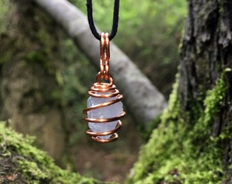 May include: A clear crystal wrapped in copper wire, hanging from a black cord. The crystal is suspended in a forest setting.