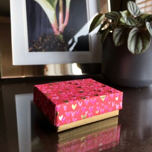 May include: A small red gift box with a gold trim and a pattern of pink hearts. The box is sitting on a dark brown surface.