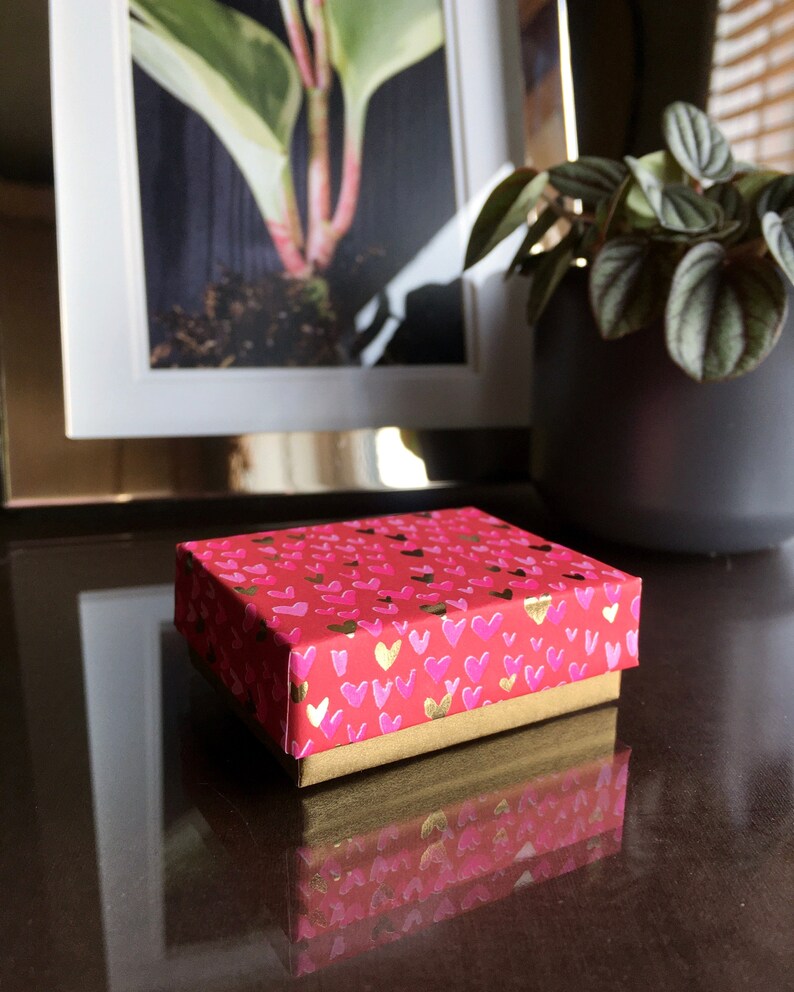 May include: A small red gift box with a gold trim and a pattern of pink hearts. The box is sitting on a dark brown surface.