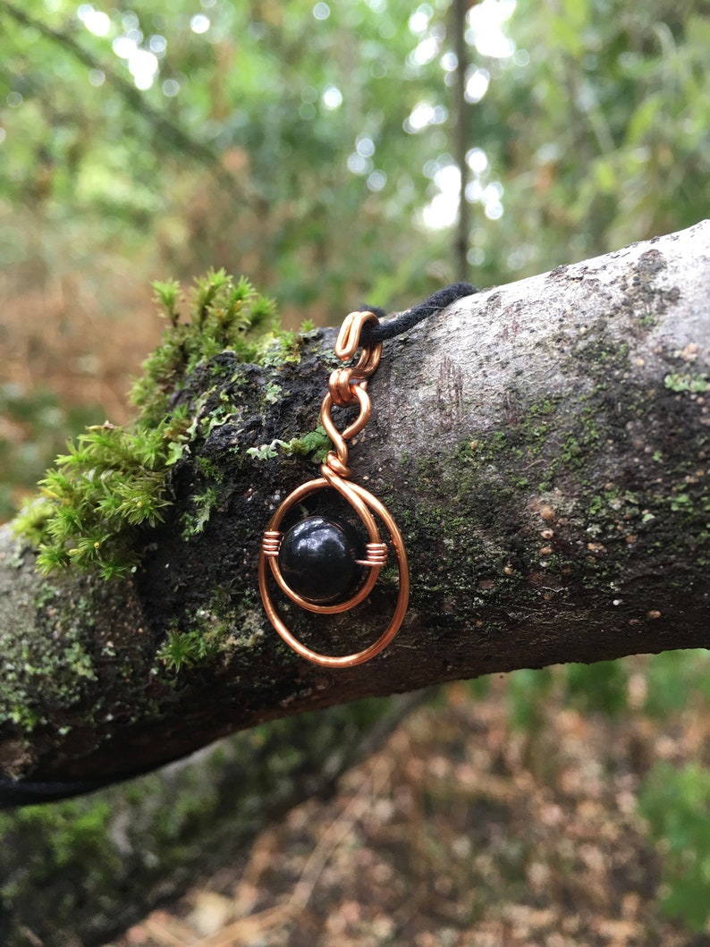 May include: A black gemstone pendant with a copper wire design. The pendant is hanging from a black cord and is resting on a tree branch covered in moss.
