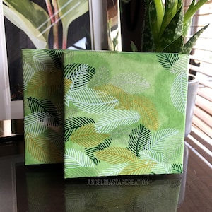 Peut inclure: Deux toiles carrées vertes avec des motifs de feuilles blanches, dorées et vert foncé. Les toiles sont exposées sur une surface réfléchissante. Le texte "ANGELINASTARCREATION" est visible en bas à droite de la toile.