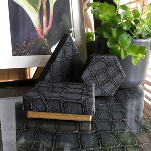 May include: Three black ring boxes with a crocodile skin pattern. The boxes are in the shape of a pyramid, a hexagon, and a square. The boxes are on a black surface with a crocodile skin pattern.