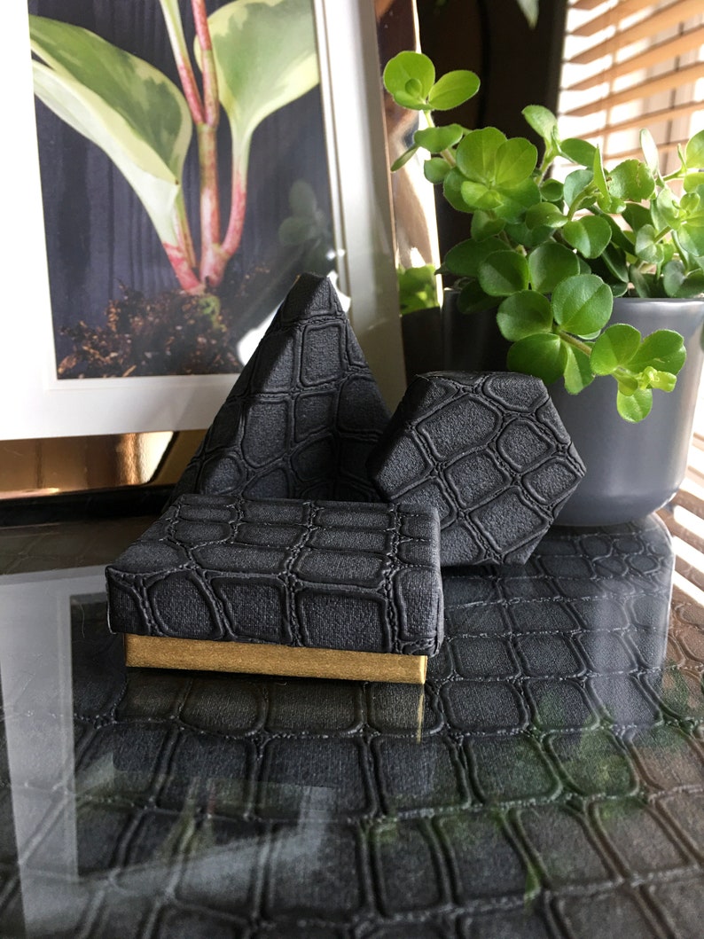 May include: Three black ring boxes with a crocodile skin pattern. The boxes are in the shape of a pyramid, a hexagon, and a square. The boxes are on a black surface with a crocodile skin pattern.