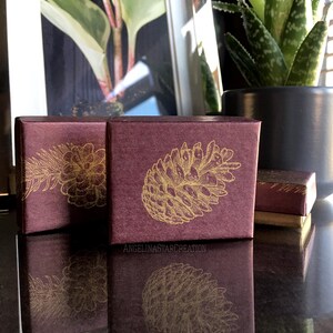 Könnte beinhalten: Burgunderfarbene Geschenkboxen mit goldenen Kiefernzapfen-Illustrationen. Die quadratischen Boxen haben eine strukturierte Oberfläche. Das Bild zeigt auch einen gerahmten Pflanzen-Druck und eine Topf-Aloe-Vera-Pflanze.
