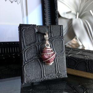 Peut inclure: Un pendentif en fil d'argent enveloppé d'une pierre précieuse de rubis rose. Le pendentif est présenté sur une surface noire texturée.