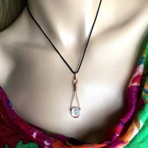 May include: A delicate necklace with a clear, round gemstone pendant. The pendant is held in a copper wire cage, creating a unique and modern look. The necklace is on a black cord.