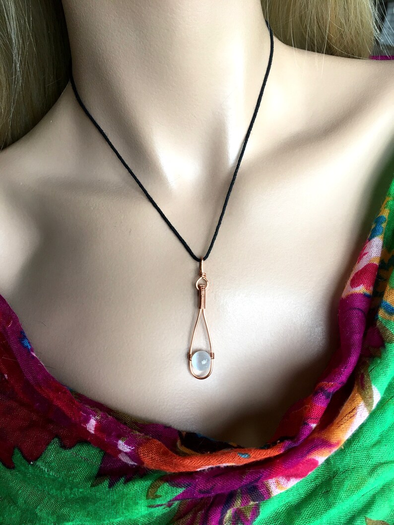 May include: A delicate necklace with a clear, round gemstone pendant. The pendant is held in a copper wire cage, creating a unique and modern look. The necklace is on a black cord.