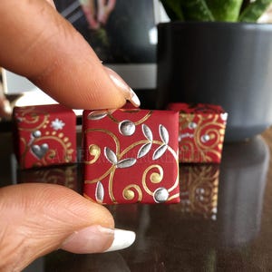 Könnte beinhalten: Nahaufnahme einer kleinen, roten quadratischen Schachtel, die mit goldenen und silbernen floralen Mustern verziert ist. Die Schachtel wird von Fingern gehalten und trägt den Text "ANGELAKARUKATION" an der Seite.