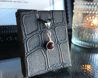 May include: A silver pendant necklace with a small, round, dark red gemstone. The pendant hangs from a black cord and is displayed on a textured, dark gray box. The box has a reptile skin pattern. The background includes a framed picture and a glass candle holder.