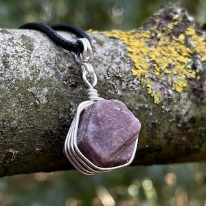 May include: A raw, deep purple-red gemstone pendant secured in a silver-coloured wire wrap. The stone has a hexagonal, multifaceted shape and is suspended from a simple black cord necklace, creating a contrast between the metal and the stone.