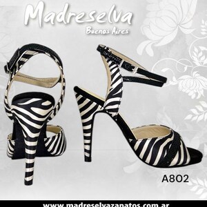 May include: Two high-heeled sandals with a zebra stripe pattern in black and white. The sandals have a thin ankle strap and a buckle closure. The heels are stiletto style. The text "Madreselva Buenos Aires" is at the top.