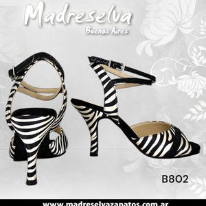 May include: Pair of black and white zebra print high-heeled sandals with ankle straps. The sandals have a buckle closure and a slender heel. The text "Madreselva Buenos Aires" is at the top, and "B802" is at the bottom.