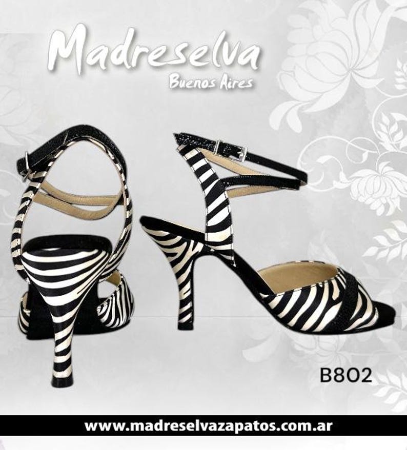 May include: Pair of black and white zebra print high-heeled sandals with ankle straps. The sandals have a buckle closure and a slender heel. The text "Madreselva Buenos Aires" is at the top, and "B802" is at the bottom.