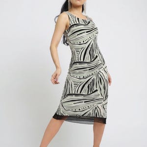 May include: Sleeveless midi dress with a black, white, and gray abstract pattern. The dress has a black mesh hem and is paired with strappy sandals. The model is standing in a studio setting.