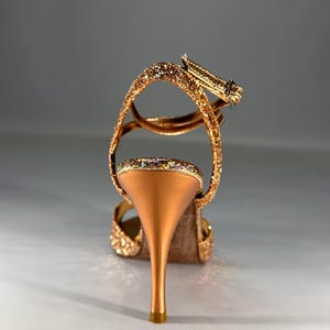 May include: A high-heeled sandal featuring a metallic copper stiletto heel and a glittery gold finish on the straps and heel cup. The shoe includes an adjustable ankle strap with a small metal buckle, showcasing a textured, sparkling surface.