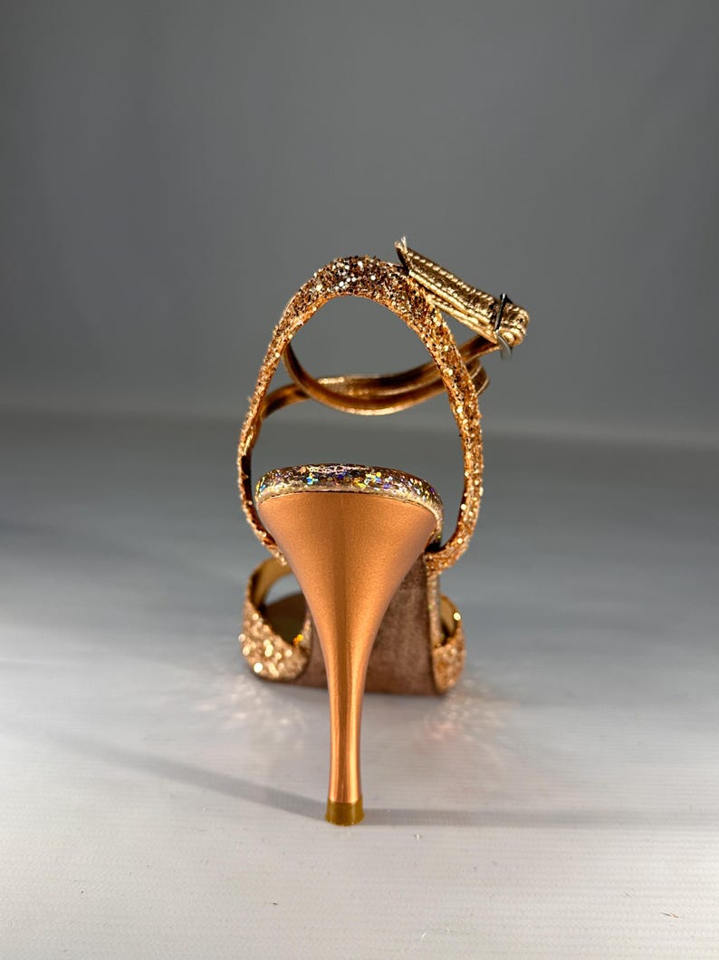 May include: A high-heeled sandal featuring a metallic copper stiletto heel and a glittery gold finish on the straps and heel cup. The shoe includes an adjustable ankle strap with a small metal buckle, showcasing a textured, sparkling surface.