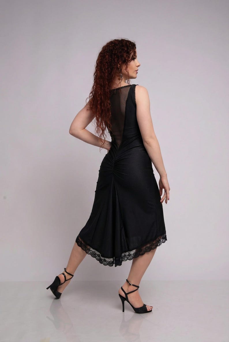 May include: Black sleeveless dress with a sheer back panel and lace trim at the hem. The dress has a fitted bodice and a flowing skirt. The model is wearing black high heels.
