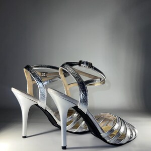May include: A pair of metallic silver high-heeled sandals featuring a multi-strap design across the vamp, a slim stiletto heel, and an adjustable ankle strap with a small silver-toned metal buckle closure. The interior sole is a neutral beige.