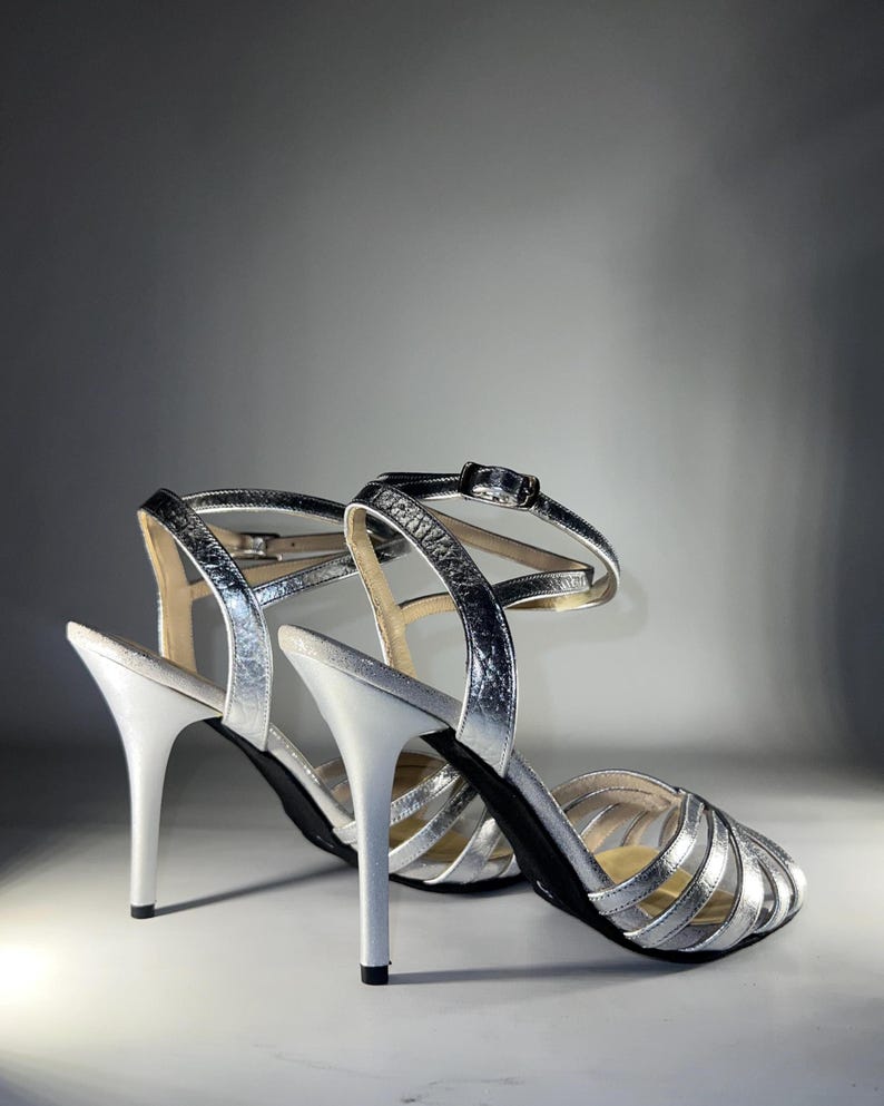 May include: A pair of metallic silver high-heeled sandals featuring a multi-strap design across the vamp, a slim stiletto heel, and an adjustable ankle strap with a small silver-toned metal buckle closure. The interior sole is a neutral beige.