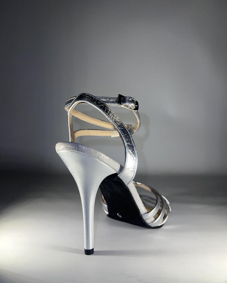 May include: A metallic silver high-heeled sandal featuring a thin stiletto heel, a black sole with the number 5 visible, and a strappy upper design with a buckle closure at the ankle. The shoe has a textured, reflective finish throughout.