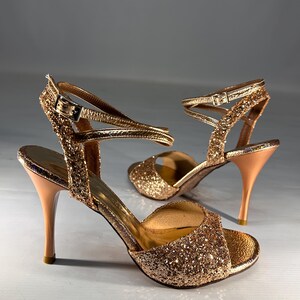 May include: A pair of gold glitter high-heeled sandals featuring an open-toe design, a slim stiletto heel, and a thin ankle strap with a metallic buckle closure. The exterior is covered in fine gold sparkles, while the interior lining is smooth.