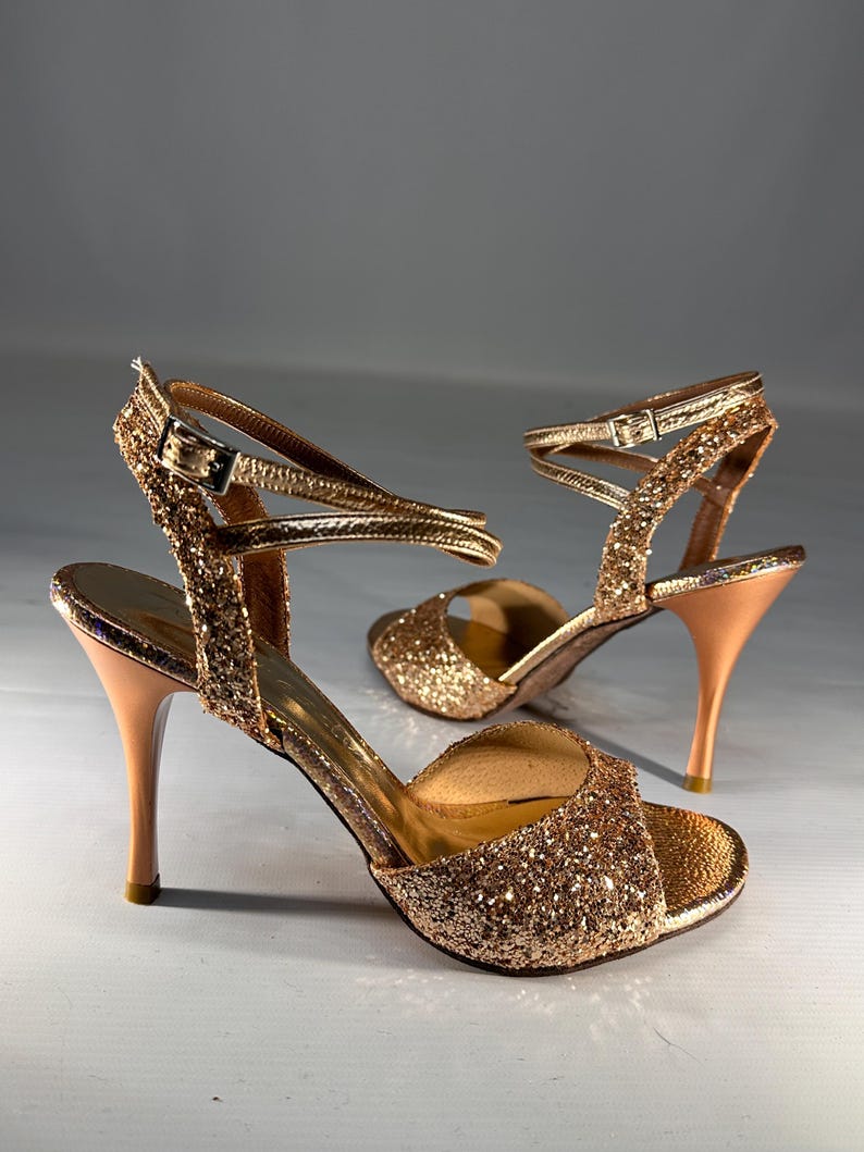 May include: A pair of gold glitter high-heeled sandals featuring an open-toe design, a slim stiletto heel, and a thin ankle strap with a metallic buckle closure. The exterior is covered in fine gold sparkles, while the interior lining is smooth.