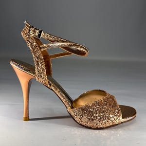 May include: A gold glitter high-heeled sandal featuring an open-toe design, a slim stiletto heel, and an adjustable ankle strap with a metallic buckle. The exterior is covered in fine gold glitter, while the heel has a smooth metallic finish.
