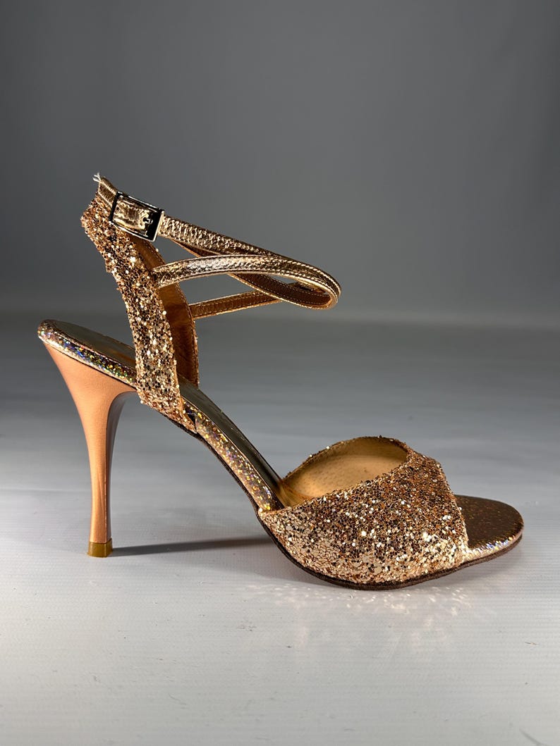 May include: A gold glitter high-heeled sandal featuring an open-toe design, a slim stiletto heel, and an adjustable ankle strap with a metallic buckle. The exterior is covered in fine gold glitter, while the heel has a smooth metallic finish.