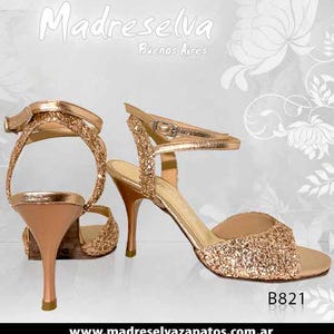 May include: A pair of rose gold glitter high-heeled sandals with ankle straps and metallic heels. The shoes feature a textured, sparkling finish on the front and heel straps, with smooth metallic material on the heel and inner sole. Text reads Madreselva Buenos Aires and B821.