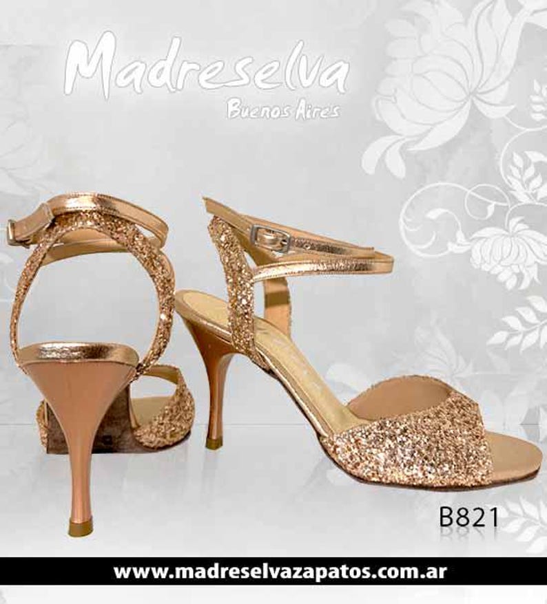 May include: A pair of rose gold glitter high-heeled sandals with ankle straps and metallic heels. The shoes feature a textured, sparkling finish on the front and heel straps, with smooth metallic material on the heel and inner sole. Text reads Madreselva Buenos Aires and B821.