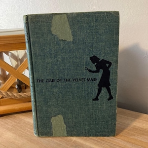 Puede incluir: Un libro antiguo titulado "The Clue of the Velvet Mask" con una cubierta verde azulado. La portada presenta una silueta negra de una persona sosteniendo una lupa. El libro está sobre una superficie de madera.