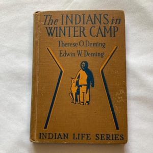 May include: A vintage book titled "The Indians in Winter Camp" with a tan cover. The cover features blue and orange illustrations of a person and a dog, with text in blue. The book is part of the "Indian Life Series".