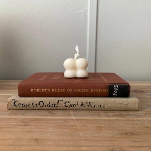May include: A small, off-white candle in a unique four-blob design sits atop a vintage, reddish-brown book titled "ROBERT'S RULES OF ORDER REVISED." Another book with the text "Come to Order!" is below.