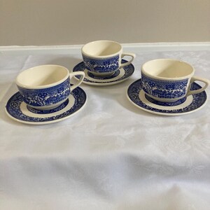 May include: Three matching teacups and saucers. The cups and saucers are white with a blue Willow pattern design. The cups have a curved handle and the saucers are round. The set is placed on a white tablecloth.